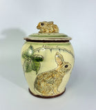 A ceramic lidded jar with a rabbit figurine on top, decorated with green leaves and thorny branches on a cream-colored glaze base.