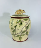 A ceramic lidded jar with a rabbit figurine on top, decorated with green leaves and thorny branches on a cream-colored glaze base.