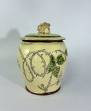 A ceramic lidded jar with a rabbit figurine on top, decorated with green leaves and thorny branches on a cream-colored glaze base.