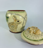 A ceramic lidded jar with a rabbit figurine on top, decorated with green leaves and thorny branches on a cream-colored glaze base.